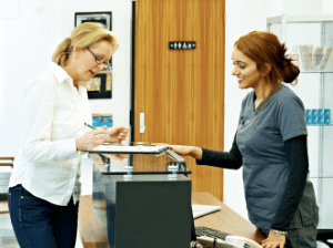 Woman signing in at a dental office reception desk while speaking with staff member.